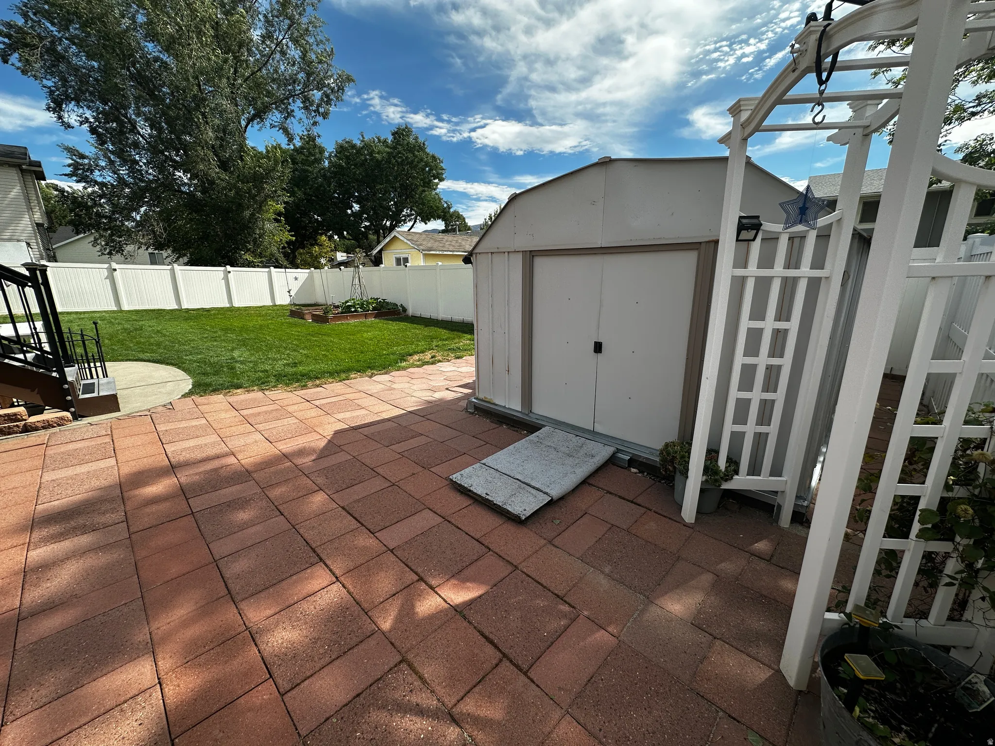 Expansive red paver patio, lush green lawn, white vinyl fencing, and a functional storage shed