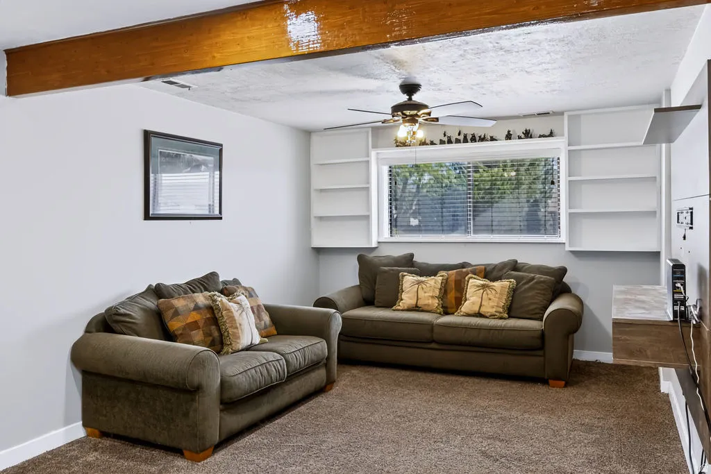 Exposed wood beam ceiling, built-in shelving, ceiling fan, window with blinds, and wall-mounted media console