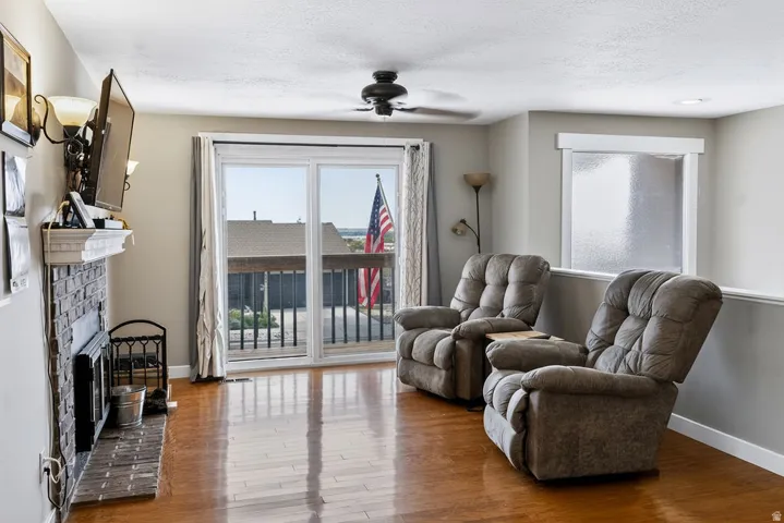 Wood-finish flooring, brick fireplace with a white mantel, sliding glass door leading to a deck with black railings, and a ceiling fan