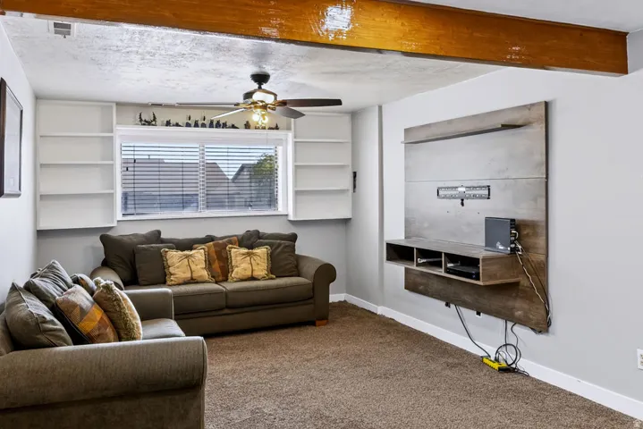 Exposed wooden beam, built-in shelving, window with blinds, ceiling fan with light fixture, and mounted media console