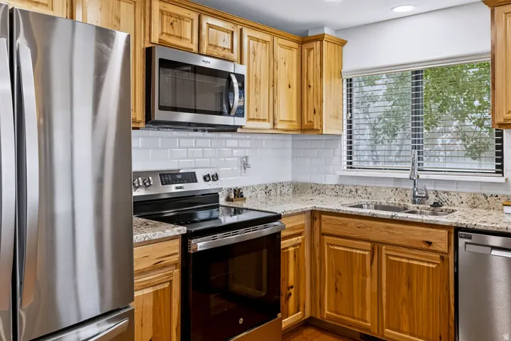 Kitchen featuring stainless steel appliances, wood cabinetry, stone countertops, white subway tile backsplash, and a double basin sink with a high-arc faucet