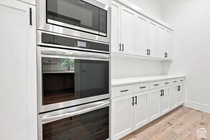 Stainless steel double oven and microwave, white shaker-style cabinetry, matte black hardware, white countertops, and wood-finish flooring