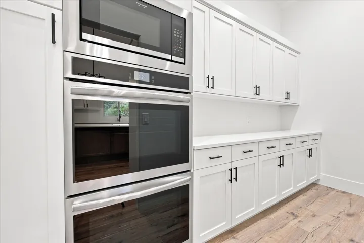 Stainless steel double oven and microwave, white shaker-style cabinetry, matte black hardware, white countertops, and wood-finish flooring