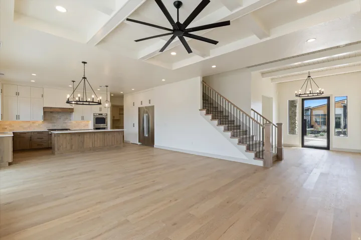 Open-concept living area featuring wood-finish flooring, a coffered ceiling with a modern ceiling fan, a kitchen island with built-in appliances, a staircase with metal balusters, and a black-framed glass entry door