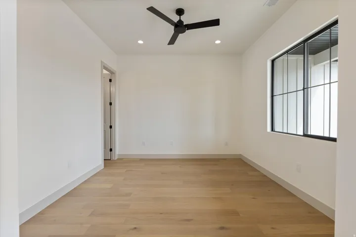 Spacious room featuring light wood-finish flooring, a contemporary black ceiling fan, recessed lighting, and a large window with black grilles