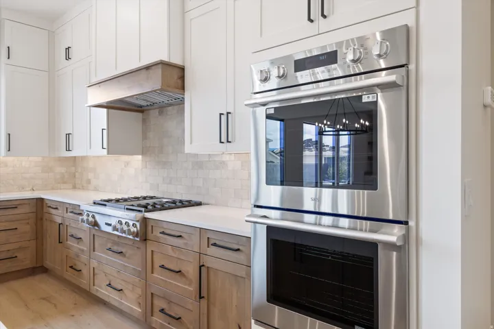 Kitchen featuring stainless steel wall oven, gas range, wood-finish lower cabinetry, white upper cabinetry, and a neutral tile backsplash