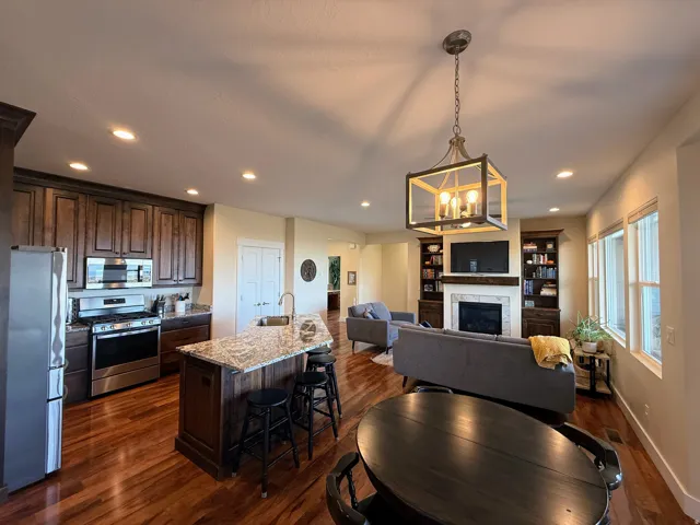 Wood-finish flooring throughout, kitchen with dark wood cabinetry and stainless steel appliances, granite island countertop with seating, living area with fireplace and built-in shelving, multiple windows providing natural light