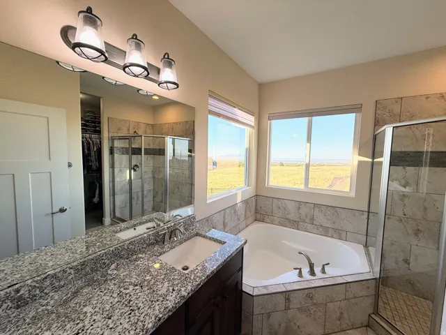Dual sink vanity with granite countertop, tiled walk-in shower with glass enclosure, corner soaking tub, and natural light from multiple windows