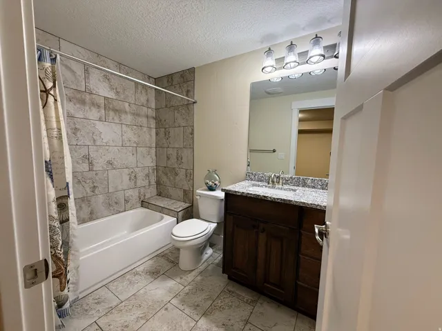 Bathtub with tiled surround, tile flooring, dark wood-finish vanity with a light-toned countertop, and a vanity mirror with attached lighting