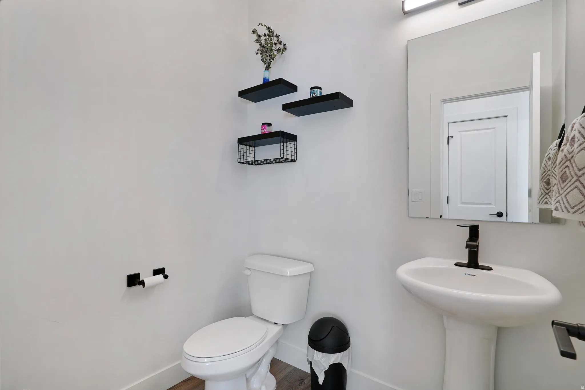 Pedestal sink with matte black faucet, wall-mounted mirror with integrated lighting, floating wall shelves, and wood-finish flooring