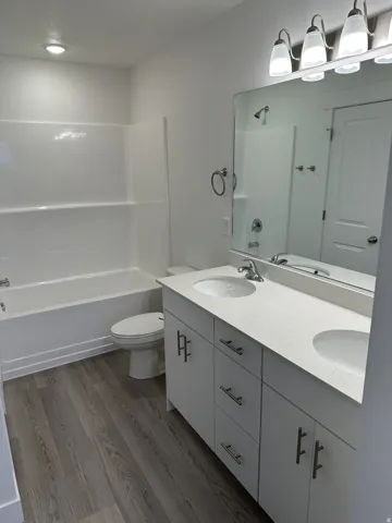 Dual vanity with integrated sinks and a light-toned countertop, wood-finish flooring, white bathtub with wall surround, and a four-light vanity fixture