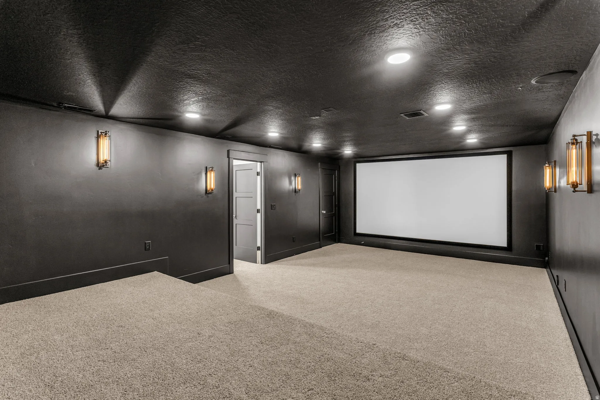 Expansive entertainment space featuring a large projection screen, recessed ceiling lighting, wall-mounted sconces, and multi-level carpeted flooring