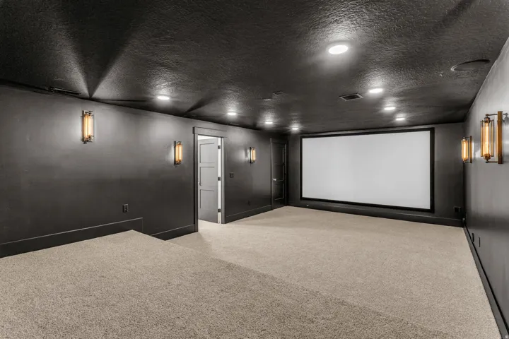 Expansive entertainment space featuring a large projection screen, recessed ceiling lighting, wall-mounted sconces, and multi-level carpeted flooring