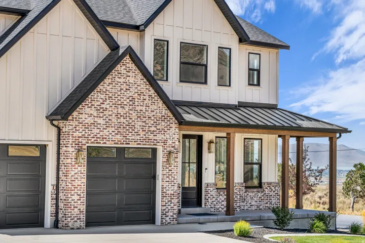 Two-story residence featuring white board-and-batten siding with brick accents, an inviting front porch with wood-finish columns, a metal roof over the porch, and dark-paneled garage doors