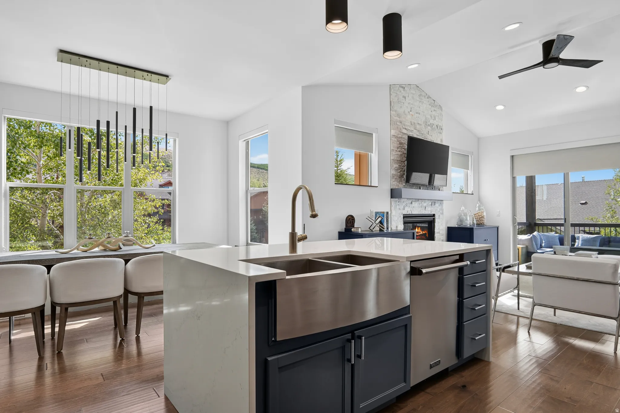 Open-concept living space with vaulted ceilings, a central kitchen island featuring a stainless steel farmhouse sink, a fireplace with a stone surround, and wood-finish flooring