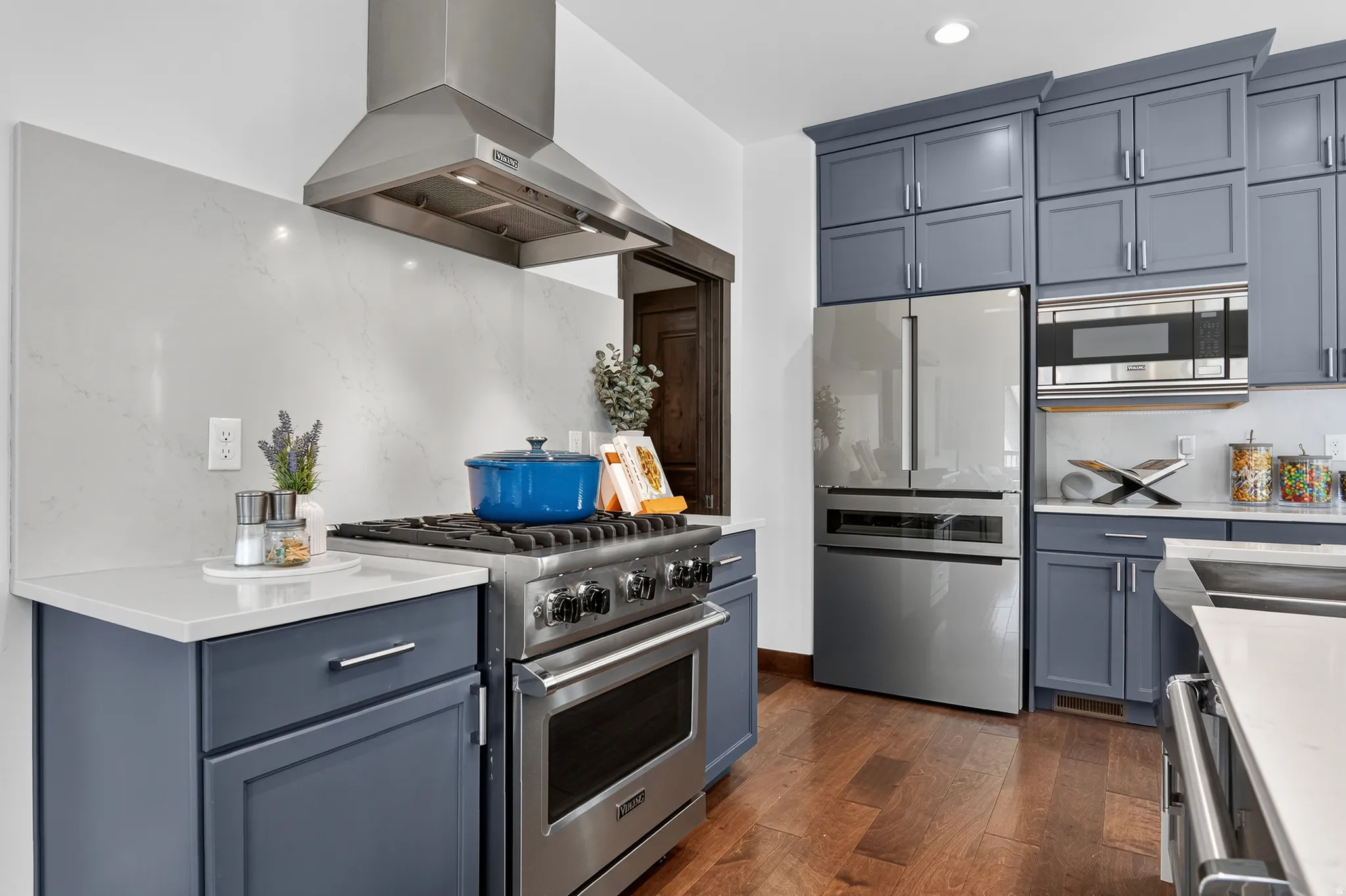 Stainless steel appliances, blue shaker-style cabinetry, white countertops, full-height backsplash, and wood-finish flooring