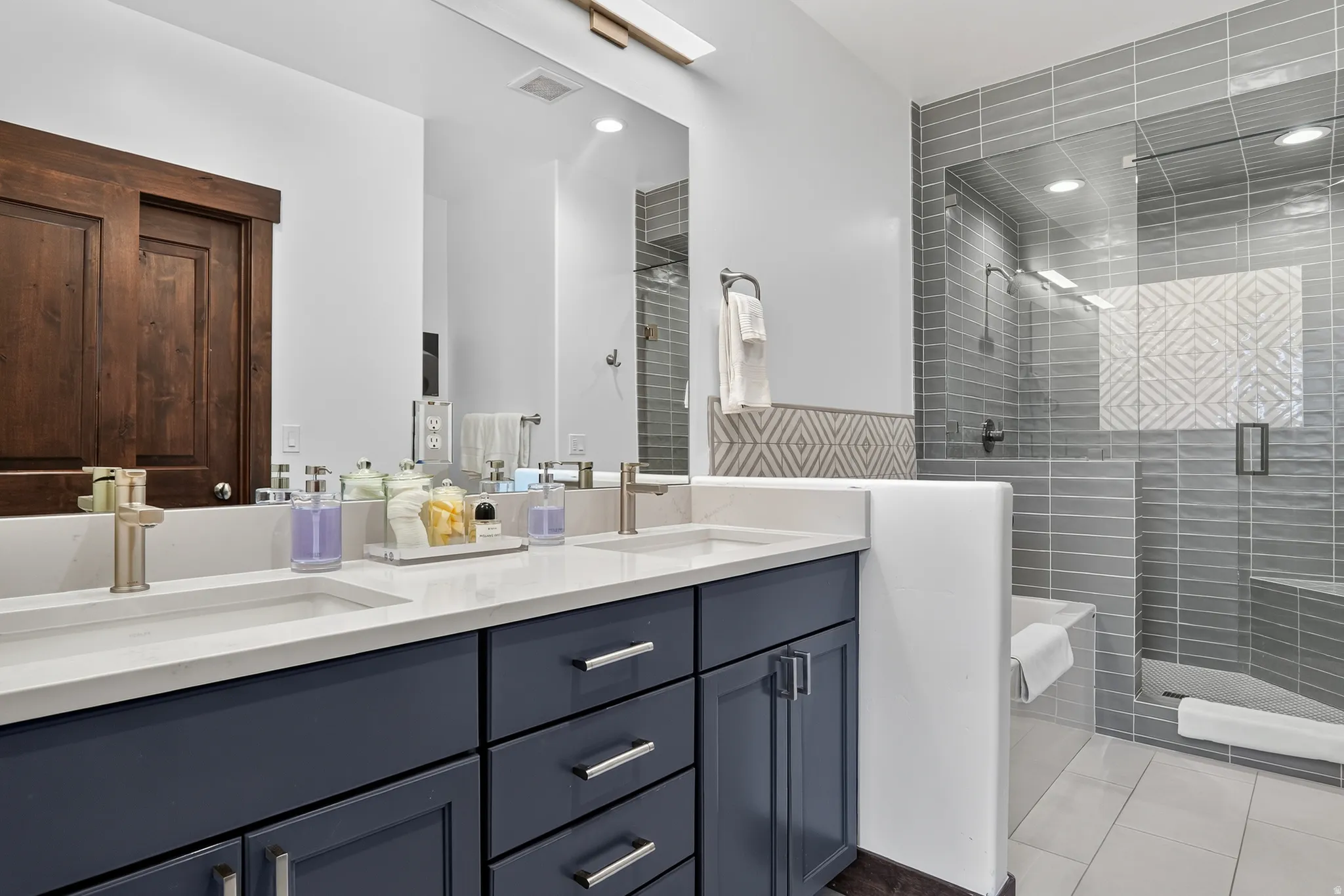 Dual sink vanity with quartz countertop, dark wood-finish barn door, glass-enclosed shower, gray subway tile, and built-in shower bench
