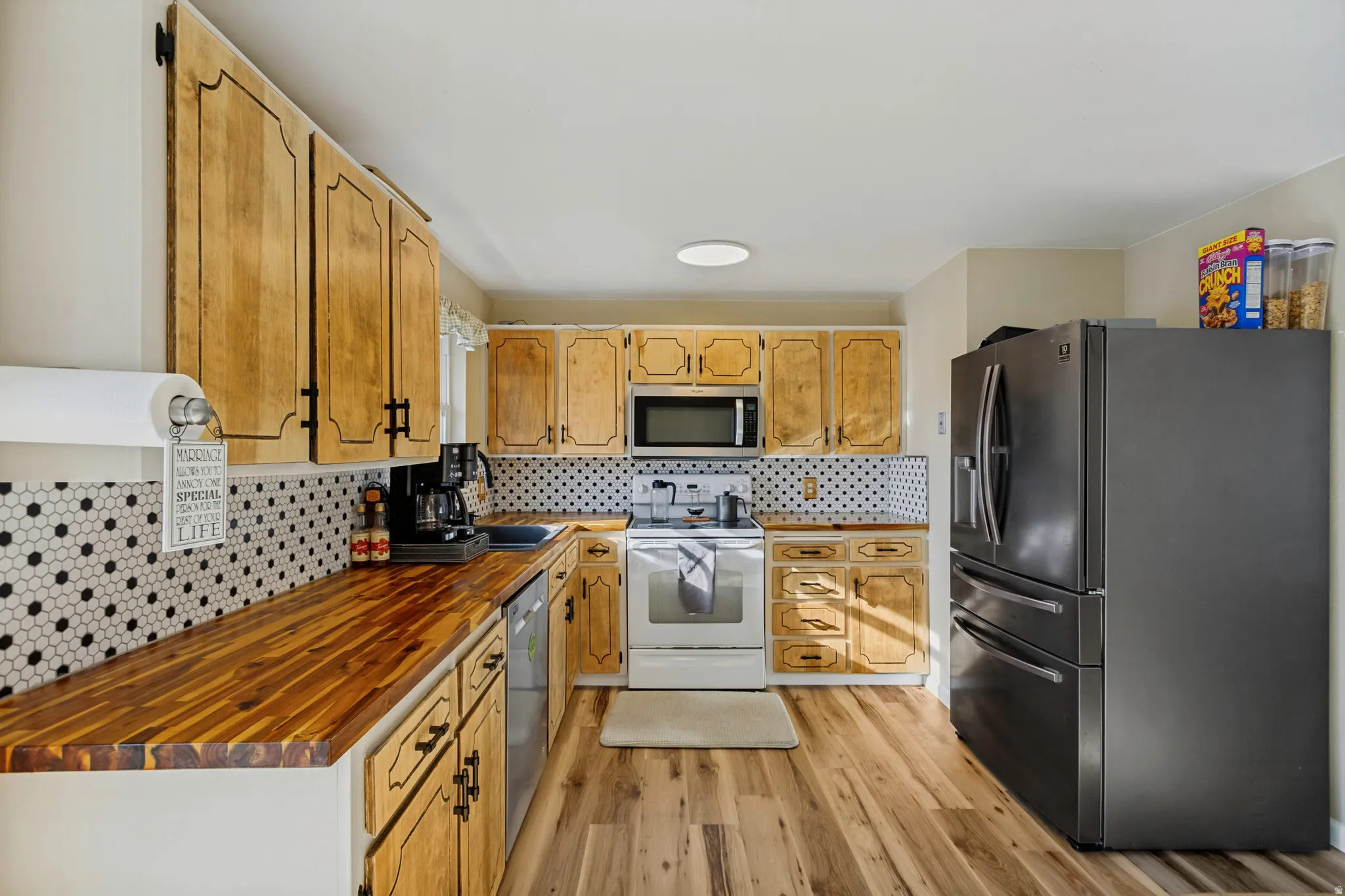 Wood-finish flooring, wood cabinetry with black hardware, hexagon tile backsplash, stainless steel appliances, and a built-in microwave