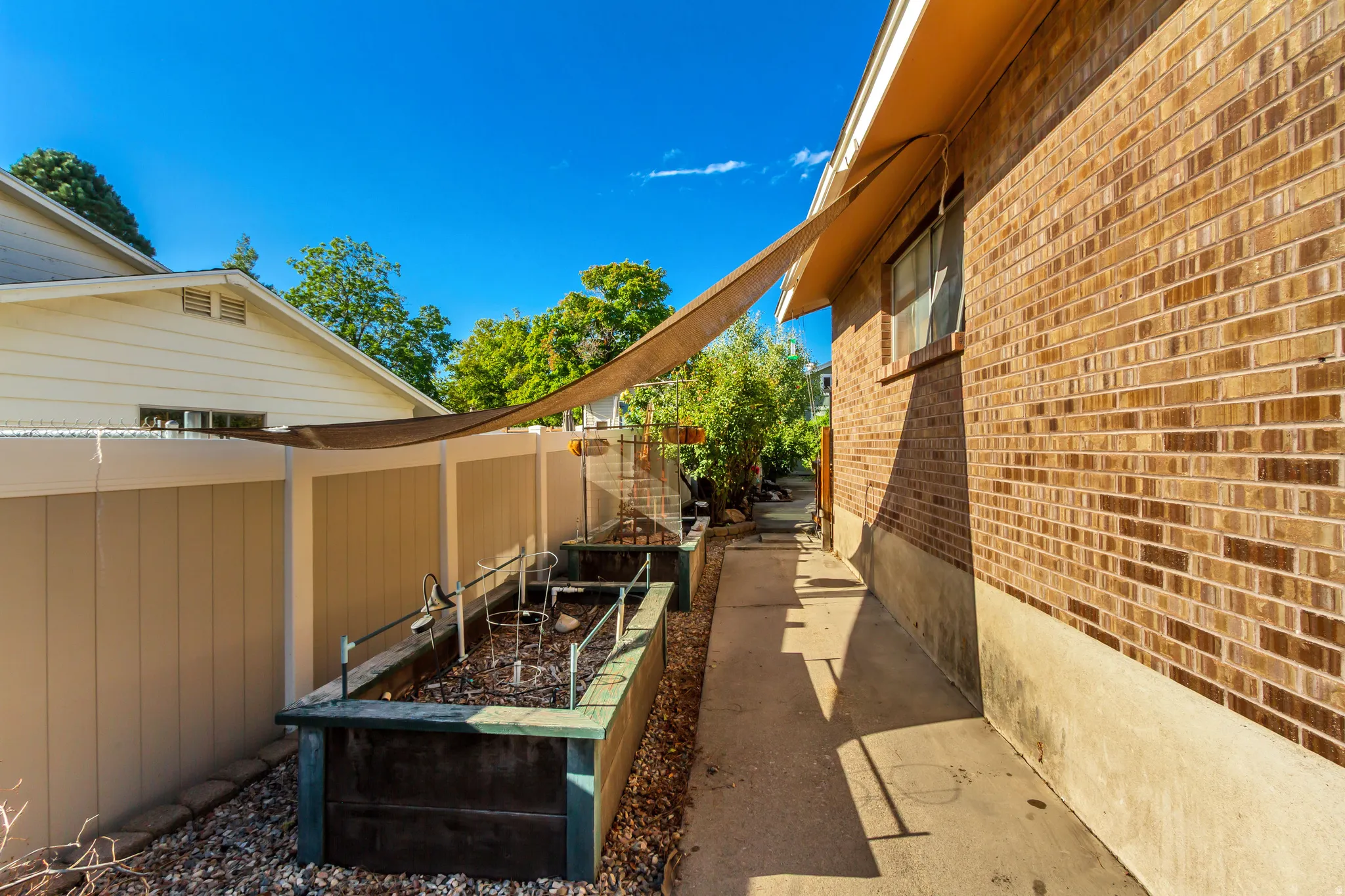 Brick exterior with a concrete walkway, raised garden beds, and a shade sail