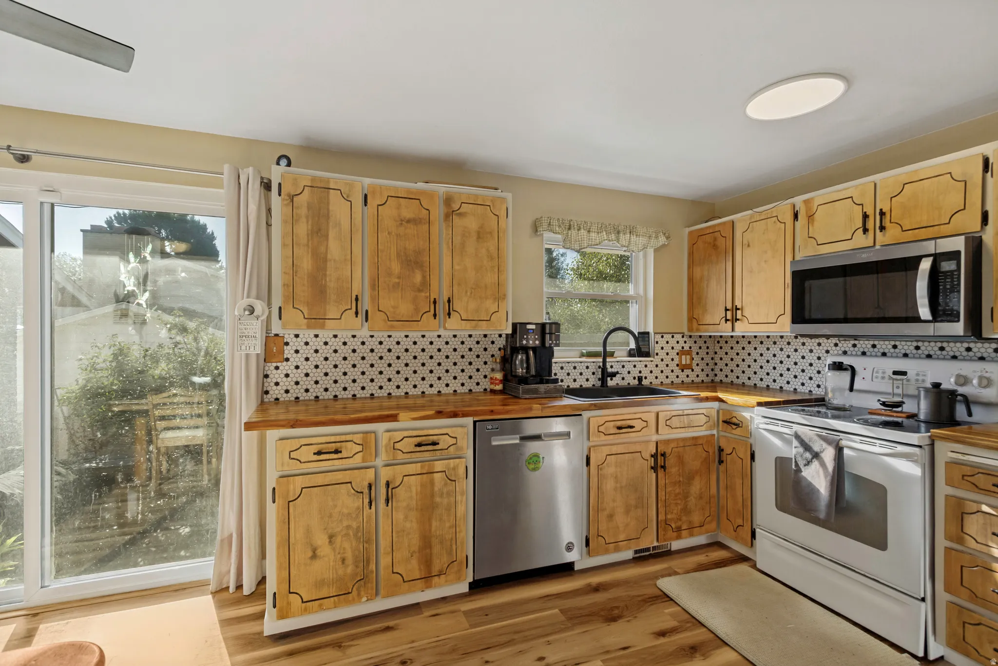 Kitchen featuring wood-finish flooring, wood cabinetry, white appliances, black and white geometric backsplash, and a sliding glass door
