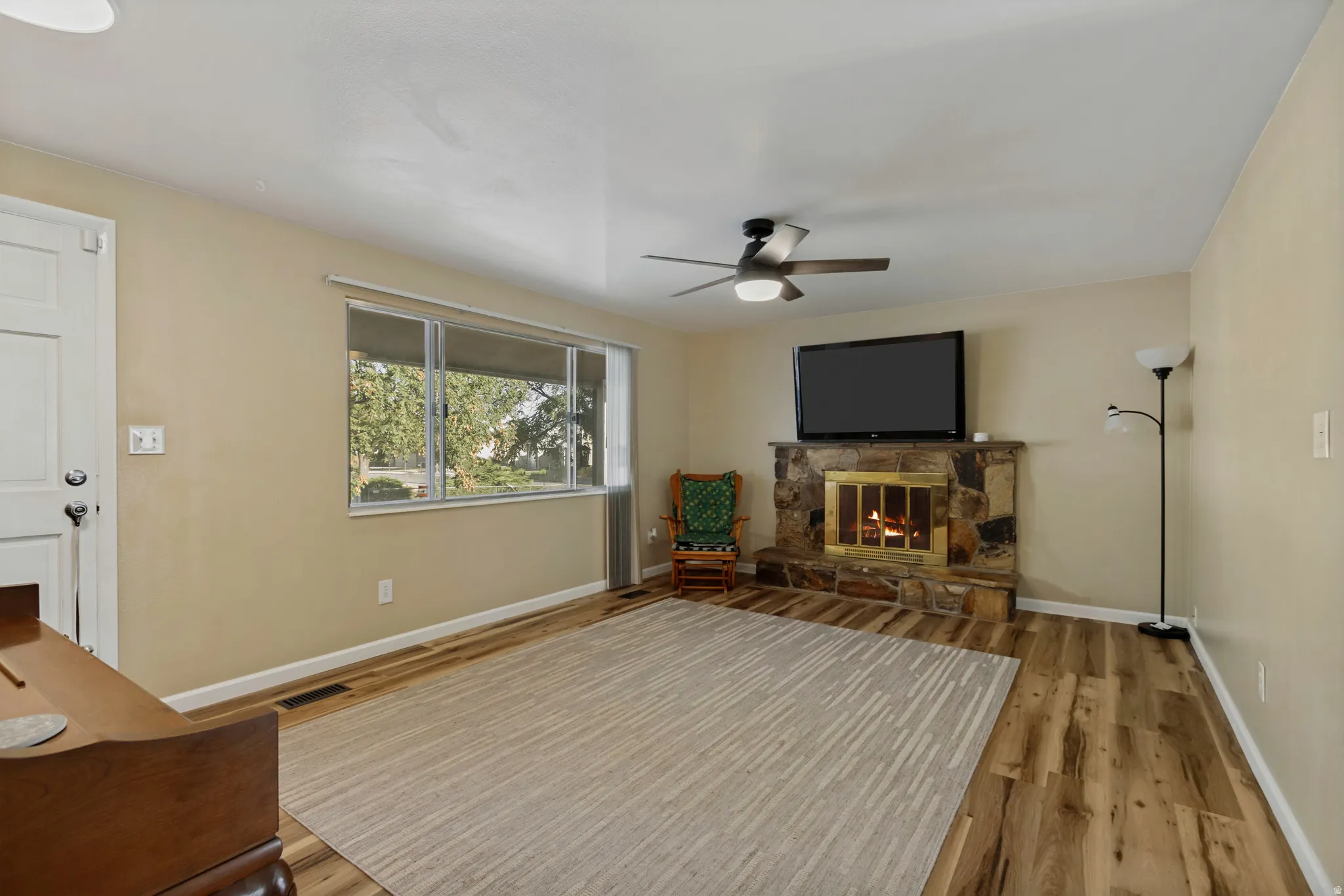 Wood-finish flooring, stone-clad fireplace with brass insert, ceiling fan with integrated lighting, and a front entry door with hardware