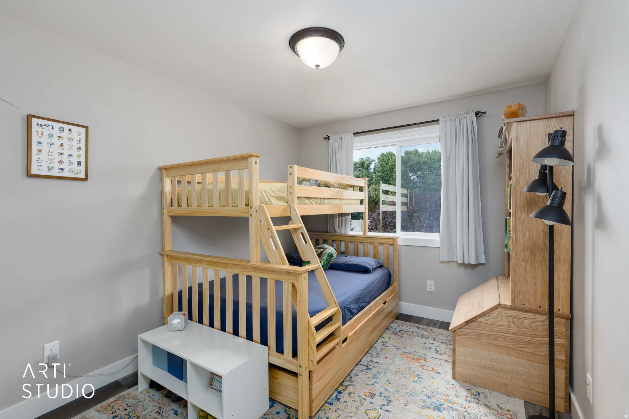 Bright, naturally lit bedroom featuring custom wood bunk bed with integrated storage, hardwood flooring, large picture window with white curtains, recessed ceiling lighting, and wood-finish wall-mounted shelving unit