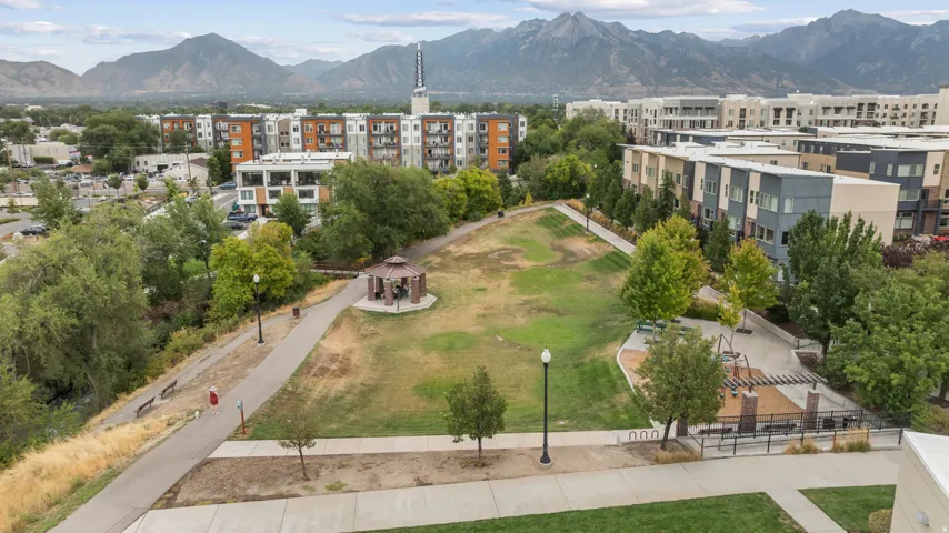 Expansive green space featuring a gazebo, walking paths, and a playground, surrounded by multi-story residential buildings and distant mountains