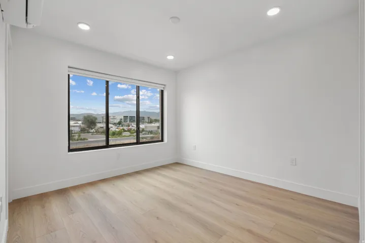 Recessed lighting, wood-finish flooring, white walls, and a large window with roller shades