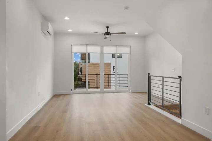 Wood-finish flooring, recessed lighting, ceiling fan, split-system AC unit, and a glass-paneled door opening to a private balcony with metal railings