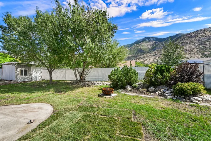 Expansive backyard featuring mature trees, a concrete patio, a storage shed with a window, a white privacy fence, and a decorative rock garden with varied plantings