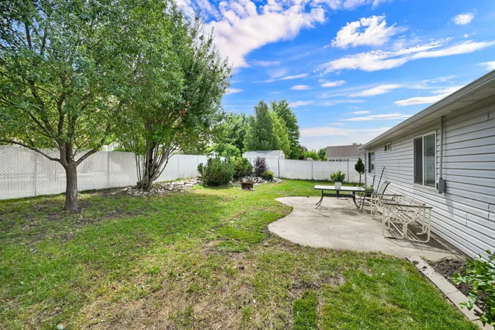 Expansive backyard featuring a concrete patio, mature trees, a white perimeter fence, and horizontal siding on the structure