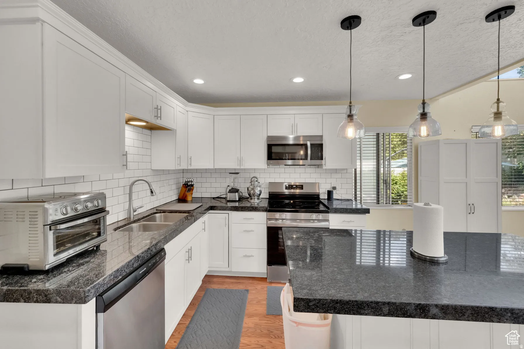 White cabinetry with brushed nickel hardware, subway tile backsplash, dark granite countertops, stainless steel appliances, and pendant lighting