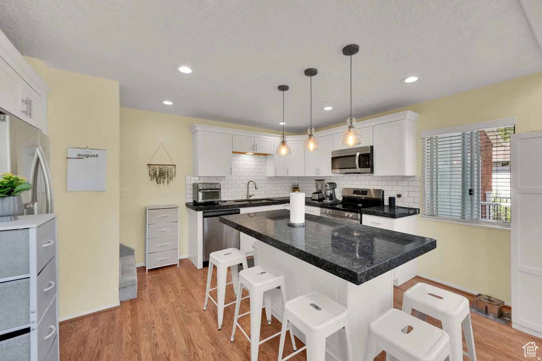 Modern kitchen featuring white cabinetry, black quartz countertops, stainless steel appliances, pendant lighting over a center island with seating for three, and warm wood-finish flooring throughout
