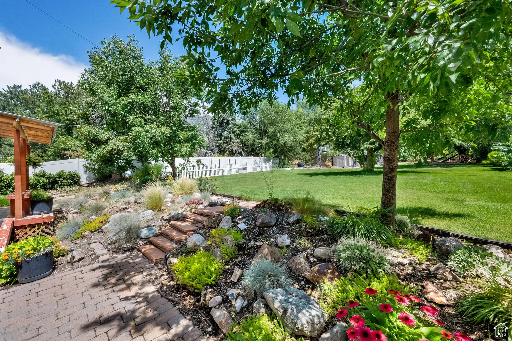Expansive backyard featuring a manicured lawn, established trees, paver patio, brick garden steps, and decorative rock landscaping