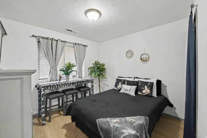 Bright secondary bedroom featuring wood-finish flooring, a large bed with black upholstered headboard, and a desk area with black checkered cabinetry positioned beneath dual windows