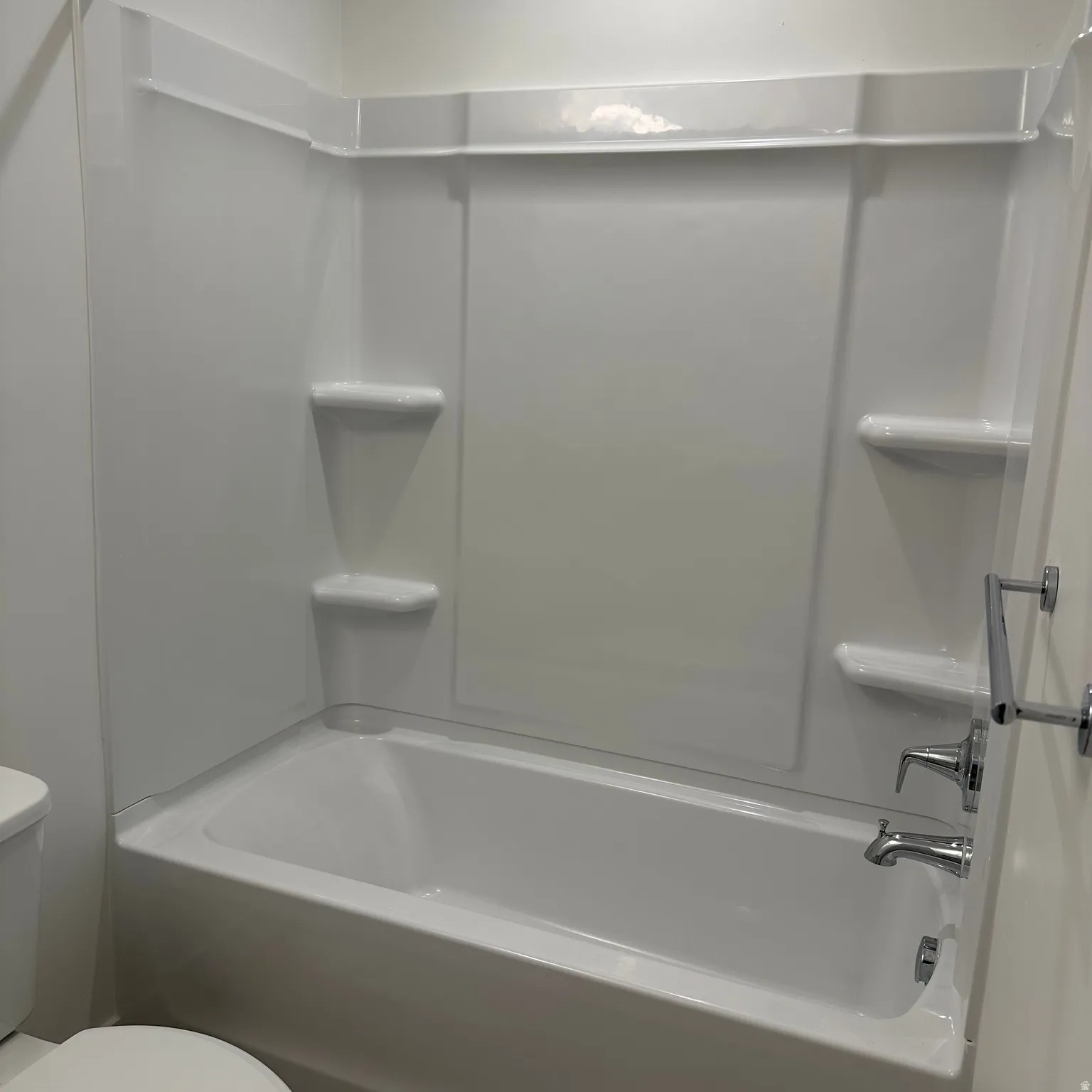 Integrated white tub and shower surround with built-in shelving, chrome fixtures, and a wall-mounted towel bar