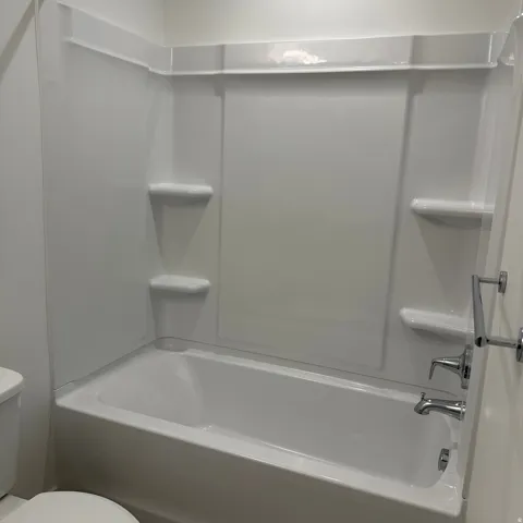 Integrated white tub and shower surround with built-in shelving, chrome fixtures, and a wall-mounted towel bar
