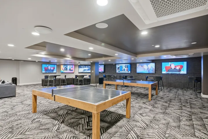 Recreation room featuring multiple built-in display screens, recessed ceiling lighting with geometric coffered details, geometric patterned flooring, and two wood-finish tables with stainless steel playing surfaces