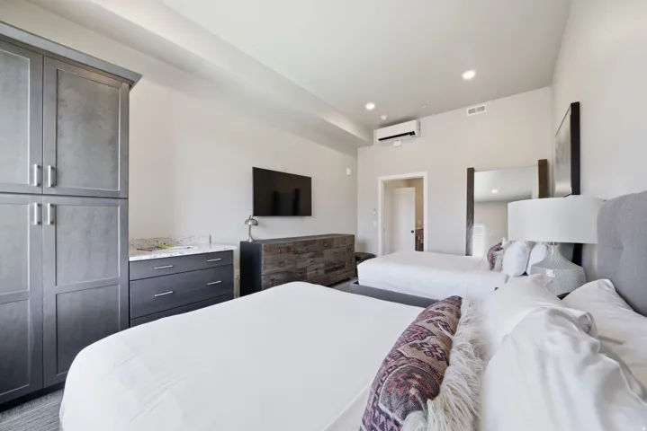 Spacious bedroom suite featuring dual queen beds with upholstered headboards, wood-finish flooring, built-in cabinetry with dark finish, wall-mounted television, and recessed ceiling lighting throughout