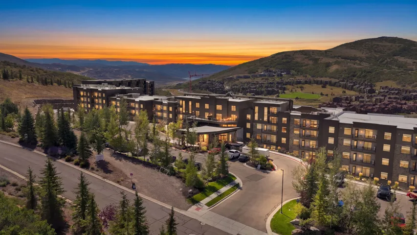 Modern multi-unit residential complex featuring contemporary architecture with illuminated facades, expansive landscaping with mature coniferous trees, professionally designed hardscaping with paved entry drives, and panoramic mountain and valley vistas a