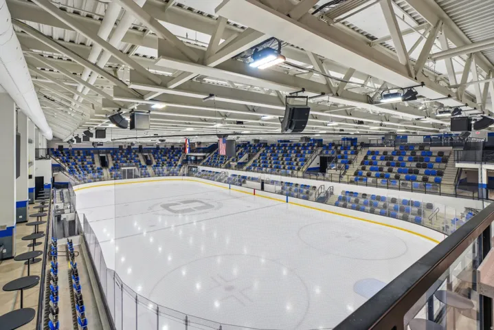 State-of-the-art ice hockey arena featuring a professional-grade playing surface with regulation boards and glass barriers, expansive seating capacity with blue upholstered seats throughout, soaring industrial ceiling with exposed structural beaming and i