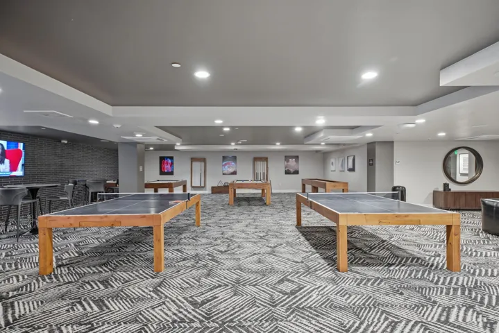 Spacious recreation area featuring geometric patterned flooring, recessed ceiling lighting, wood-finish tables, built-in bar seating with dark cabinetry, and a wall-mounted digital display