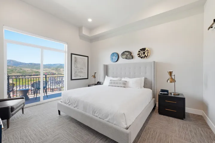 Bright primary bedroom featuring floor-to-ceiling glass doors opening to a private balcony with mountain vistas, a upholstered bed frame with vertical paneling detail, wood-finish flooring, a black nightstand with brass hardware, and three decorative wall