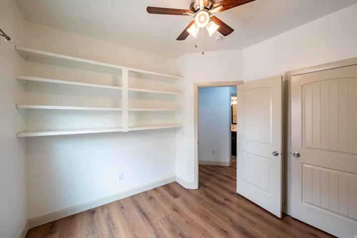 Versatile bedroom or office space featuring custom built-in shelving with adjustable shelves, wood-finish flooring, ceiling fan with integrated lighting, and dual closets with panel doors
