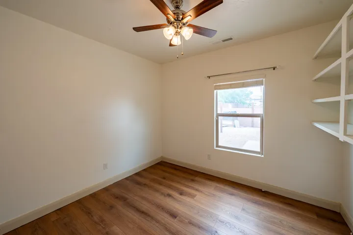 Wood-finish flooring, ceiling fan with light fixture, built-in shelving, and window with roller shade