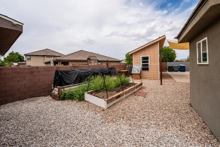 Gravel backyard featuring a storage shed with a lean-to roof, multiple raised garden beds, a concrete patio area, and a privacy wall