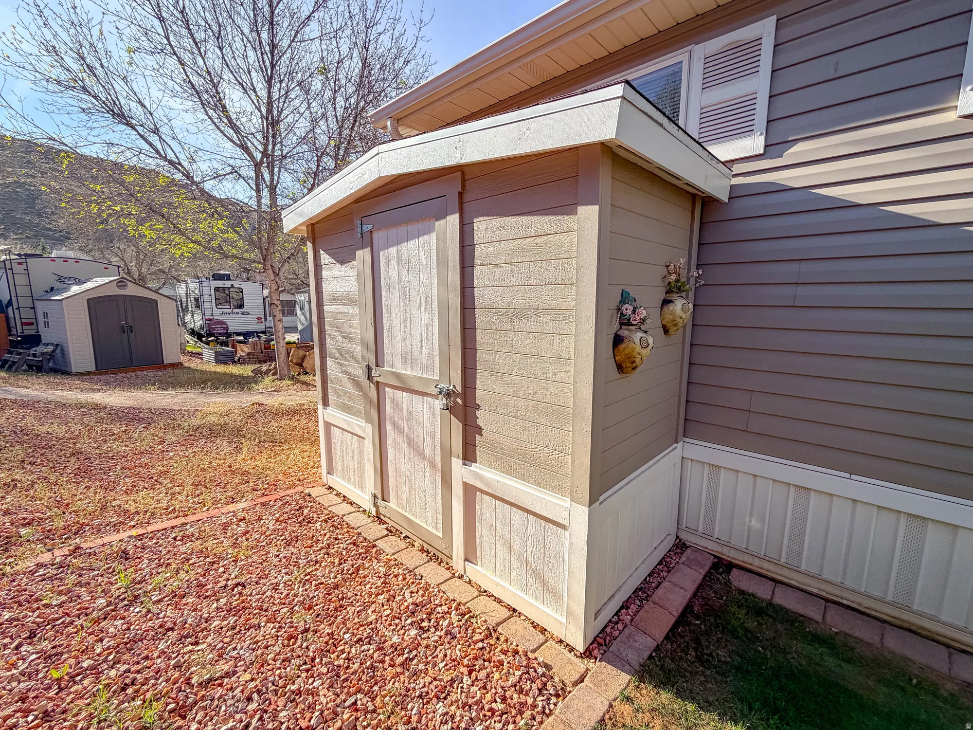 Charming detached storage structure featuring a peaked roof with wood-finish overhang, cream-colored vertical plank siding, and double hinged doors