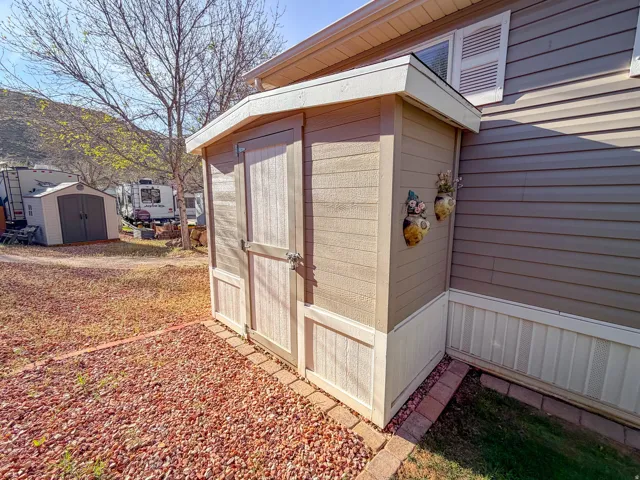 Charming detached storage structure featuring a peaked roof with wood-finish overhang, cream-colored vertical plank siding, and double hinged doors