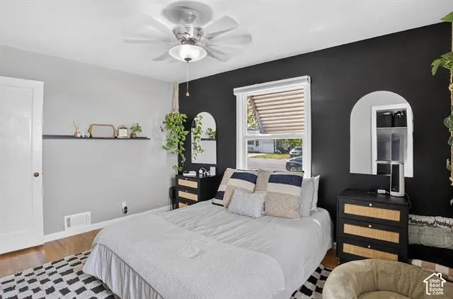 Primary Bedroom featuring a ceiling fan with integrated lighting, black accent wall with arched mirror detail, built-in wood-finish dresser with drawer storage, large window with horizontal blinds, and geometric patterned flooring