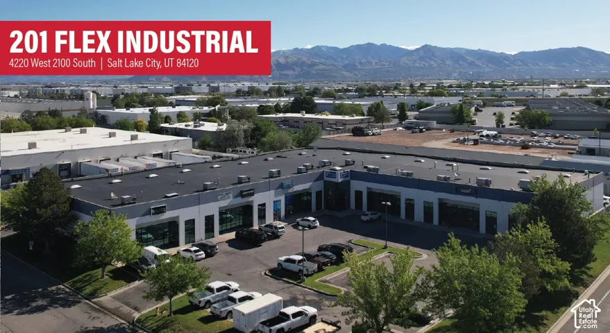 L-shaped industrial facility featuring a flat roof with multiple HVAC units, expansive paved surface parking, glass storefront entryways, and mountain scenery